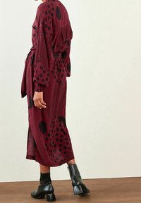 Abito borgogna con pois neri, maniche lunghe, vita con cintura e spacchi laterali, abbinato a stivali neri fino alla caviglia e ambientato su uno sfondo chiaro.
