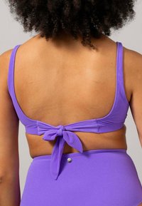 Lila bikiniöverdel med en bred band och justerbara axelremmar, med en knytdel bak. Slät textur med minimala hårdvaru detaljer.