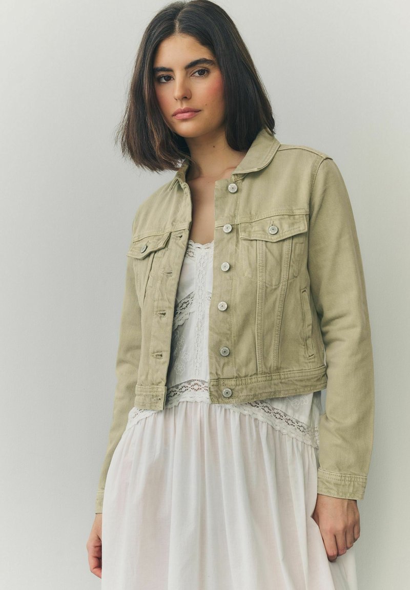 Khaki denim jacket with a collar, front pockets, and silver buttons, worn over a white lace-trimmed dress. Soft texture and fitted design.