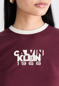 Maroon cotton shirt with white crew neck and raised logo "CALVIN KLEIN 1968" in bold font at the center. Smooth texture, standard fit.