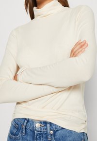 Crème-kleurige coltrui gemaakt van zachte, rekbare stof. Heeft lange mouwen en een aansluitende snit, gecombineerd met blauwe jeans.