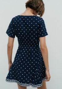 Marinblå klänning med vita prickar, korta ärmar och elastisk midja. Har rund halsringning och en vågig fåll.