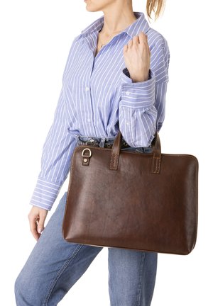 Donna con camicia a righe blu e jeans che tiene una grande valigetta di pelle marrone per i manici su sfondo bianco.