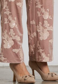Gambe inferiori che indossano pantaloni di velluto rosa con motivi floreali di paillettes e décolleté in suede beige con fiocchi decorativi.