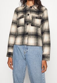 Koszulowa kurtka w kratę w odcieniach beżu i czerni, wykonana z teksturowanej tkaniny, z dwoma przednimi kieszeniami i czarnymi guzikami. Noszona na niebieskich dżinsach.