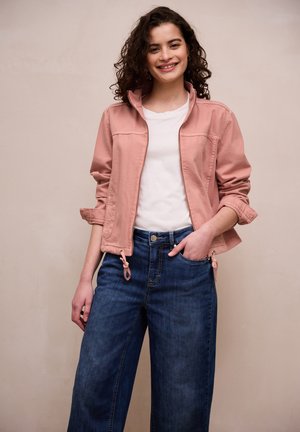 Jonge vrouw met krullend haar, gekleed in een roze jas, wit T-shirt en blauwe spijkerbroek, glimlachend met één hand in haar zak.