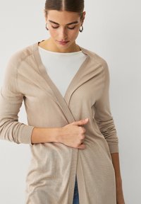Cardigan beige chiaro con colletto a scialle e design aperto, caratterizzato da polsini e orlo a coste. Indossato sopra una camicia bianca.