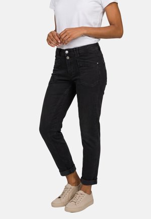 Vrouw draagt opgerolde zwarte jeans, wit T-shirt en beige sneakers, staat met handen voor zich gevouwen tegen een eenvoudige achtergrond.