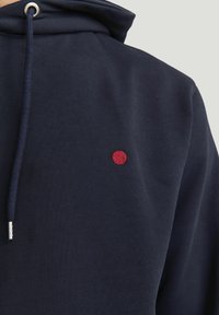 Sweatshirt marine en coton doux, doté d'un cordon de serrage et d'un petit point rouge brodé sur la poitrine. Texture lisse avec un design minimal.
