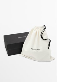 Sac en coton à cordon de couleur crème avec "Massimo Dutti" imprimé, à côté d'une boîte rectangulaire noire présentant le même logo en haut.