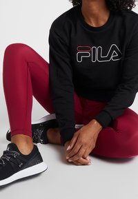 Felpa nera con logo FILA, leggings bordeaux e scarpe da ginnastica nere. I leggings hanno una texture liscia; le sneakers hanno una finitura testurizzata.