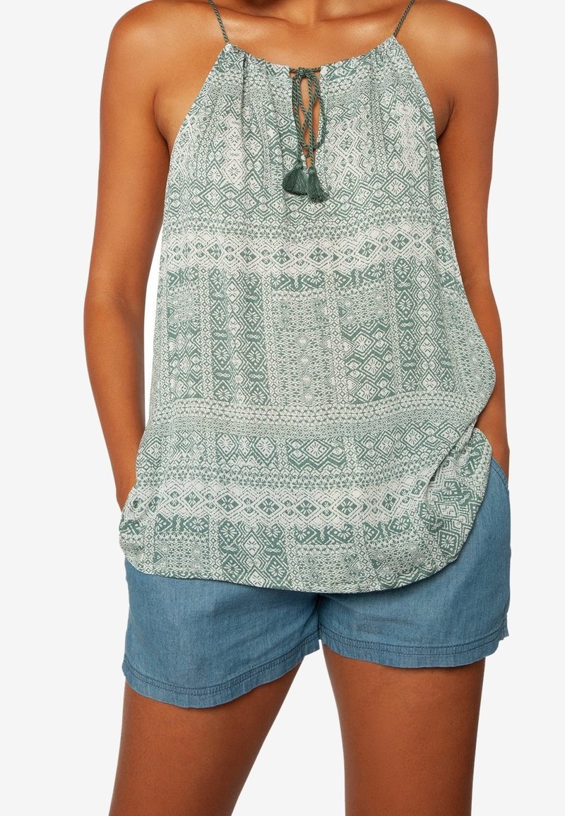 Femme portant un haut dos nu vert à motifs avec des liens à glands et un short en jean bleu, mains dans les poches.