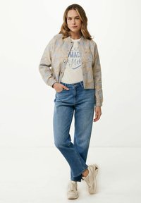 Veste bombardier bleu clair avec des motifs floraux dorés, t-shirt graphique crème, jeans bleu clair et baskets beiges. Tenue décontractée.