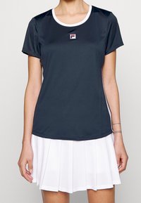 Tmavě šedý sportovní top s krátkým rukávem a bílými akcenty, na němž je logo Fila. V kombinaci s bílou skládací sukní, navrženou pro sportovní aktivity.