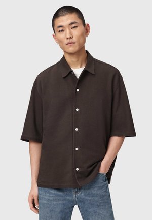 Ung mand med kort hår iført en mørkebrun kortærmet knap-op-skjorte over en hvid undertrøje og blå jeans, stående med den ene hånd i lommen.