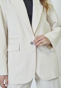 White blazer with a single black button, notch lapels, and side pockets. Smooth fabric with a tailored fit, featuring a clean, minimalist design.