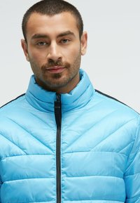 Light blue puffer jacket with a high collar, zip front, and chevron panel design. Smooth texture with black accents on shoulders.
