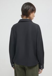 Schwarzes Langarmshirt mit einem Umlegekragen, glatter Textur und geknöpften Manschetten. Faltenfreier Stoff fällt sanft über die Schultern.