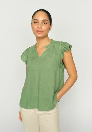 Mujer con el cabello recogido, que lleva una blusa sin mangas de volantes color verde claro y pantalones beige, de pie frente a un fondo liso y claro.