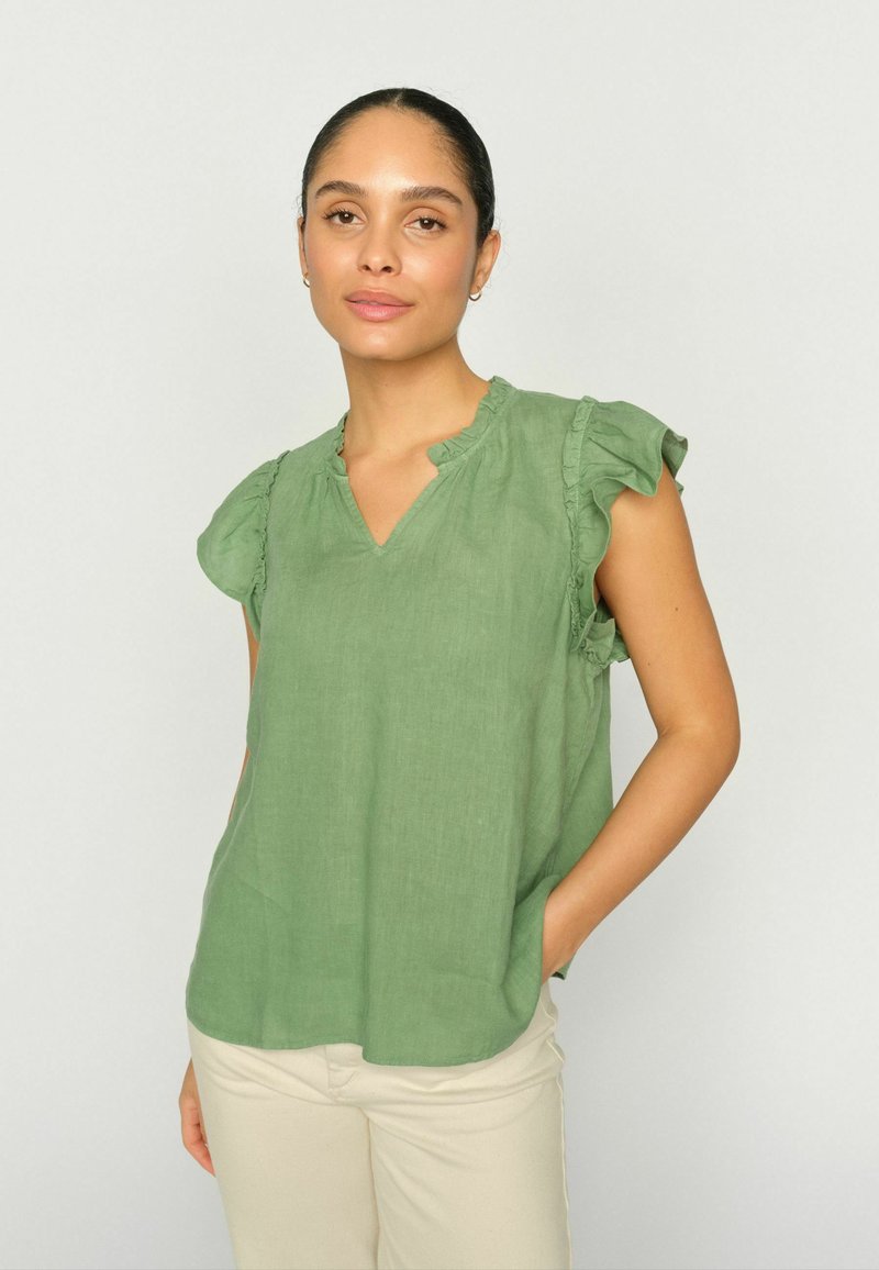 Naine, kellel on seljatatud juuksed, kannab helerohelist volangidega varrukateta pluusi ja beeže pükse, seisab ühetaolise heleda tausta ees.