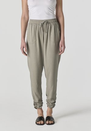 Mujer con pantalones de cintura elástica y cordón ajustable de color gris claro con bolsillos y sandalias negras sin cierre, de pie contra un fondo liso.