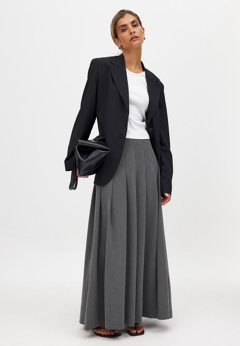 Schwarzer übergroßer Blazer über einem weißen T-Shirt, kombiniert mit einem langen grauen Faltenrock. Das Model hält eine schwarze Handtasche. Schlichte Sandalen runden den Look ab.