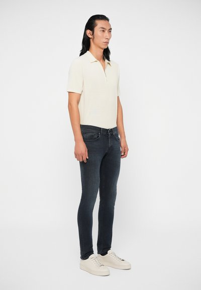 Άνδρας με μακριά μαύρα μαλλιά, ντυμένος με κρεμ πόλο κοντομάνικο, σκ ούρες στενές τζιν και άσπρα αθλητικά παπούτσια, που στέκεται σε λευκό φόντο.