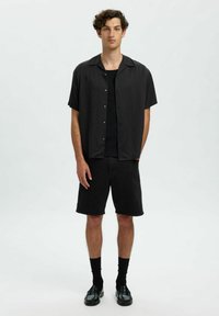Melnas īso piedurkņu krekls ar pogām un brīvu piegriezumu, apvienots ar melnām šortiem. Modelis valkā melnas īsās zeķes un melnus loafers.