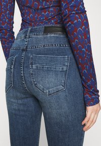 Jeans ajustados en denim azul oscuro con un acabado desvaído. Presenta dos bolsillos traseros y un parche de cuero negro de la marca en la cintura.