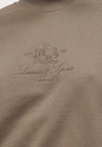 Μπεζ πουλόβερ με λαιμόκοψη ζιβάγκο, κεντημένο με έμβλημα και κείμενο "Leisure & Sports New York Estd 1926" στο στήθος.