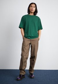 Grøn oversized bomulds t-shirt med et subtilt logo, parret med brune lige bukser og lilla sko med sorte accenter.