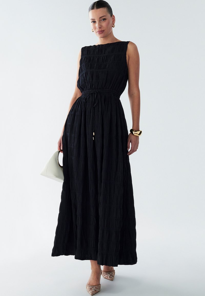 Schwarzes Maxikleid aus strukturiertem Stoff, ärmelloses Design und taillierter Schnitt. Kombiniert mit einer weißen Tasche und verzierten Spitzenschuhen.