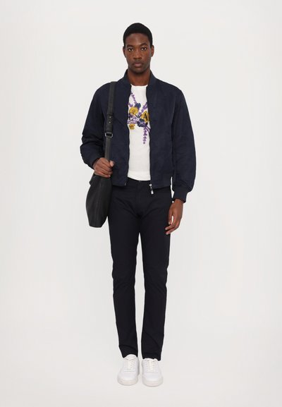 Navy bomber jacket, white graphic t-shirt, dark pants, and white sneakers. Black shoulder bag with a smooth texture.