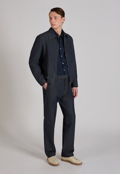 Dark denim jacket with contrasting stitching, paired with a navy button-up shirt and matching dark trousers. Beige sneakers complete the outfit.