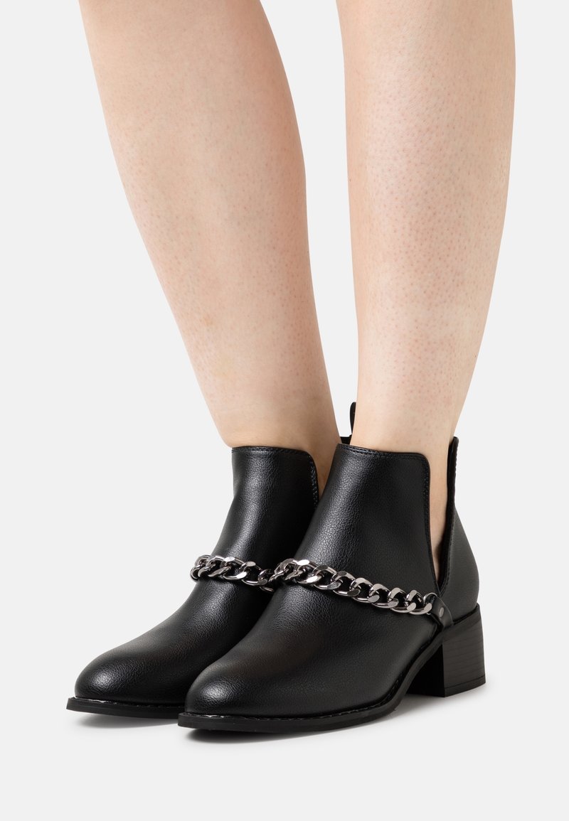 Bottines noires à talons blocs bas avec détail de chaîne argentée portées sur jambes nues sur un fond uni.