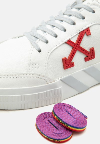 Άσπρο δερμάτινο αθλητικό παπούτσι με κόκκινο λογότυπο διπλού βέλους και γκρι κορδόνια, συνοδευόμενο από δύο κυλινδρισμένα μοβ κορδόνια με πολύχρωμα περιθώρια.