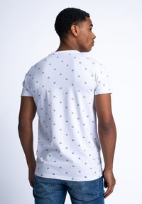 Tricou alb din bumbac, cu un model repetitiv de soare verde, cu mâneci scurte și decolteu rotund, purtat cu blugi din denim albastru.