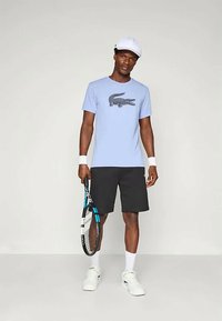 Lyseblå t-skjorte med svart krokodillelogo, kombinert med svarte shorts. Holder en tenniskølle, har på seg hvit cap og håndleddbånd. Hvite joggesko.