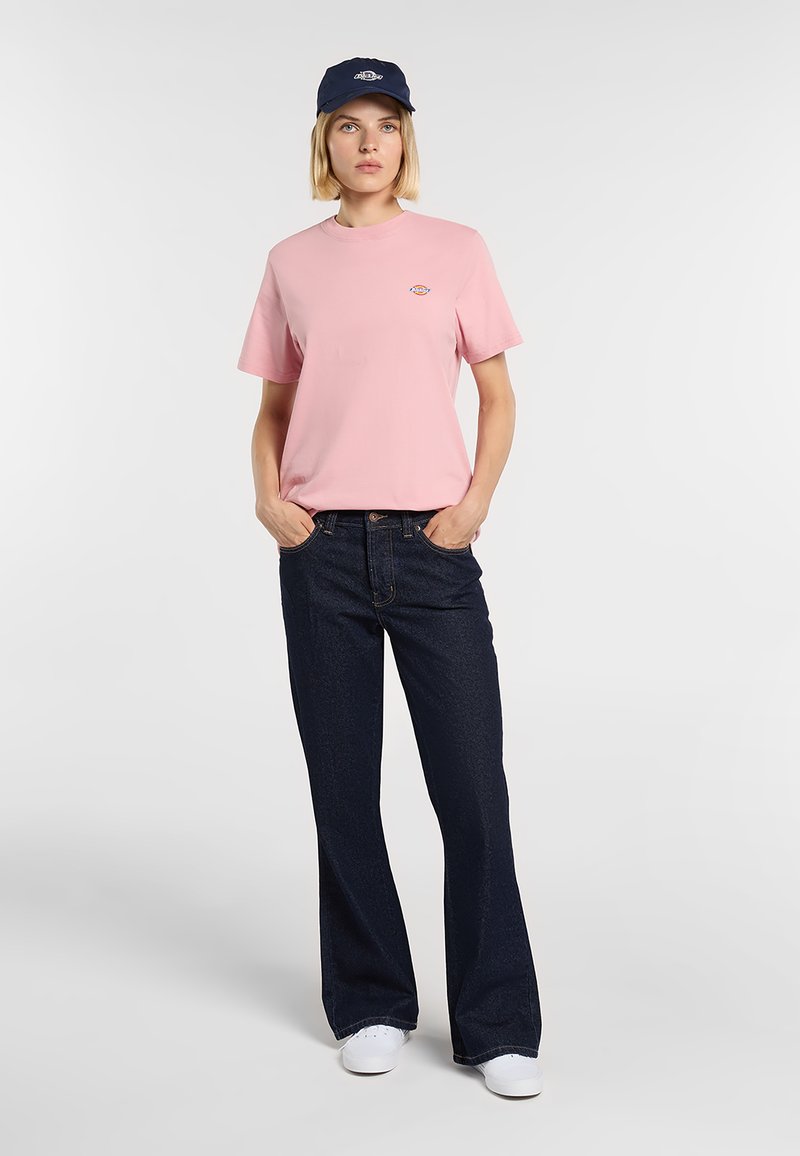 Lichtroze katoenen T-shirt met korte mouwen, gecombineerd met donkerblauwe denim flared broek en een marineblauwe pet met een logo. Witte sneakers.