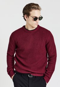 Junger Mann mit runden schwarzen Sonnenbrillen, einem strukturierten kastanienbraunen Pullover und schwarzen Hosen, die Hände in den Taschen, der seitlich schaut.