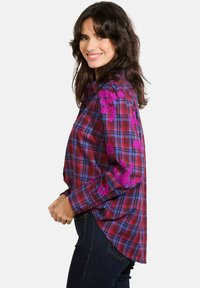 Hedvábná košile s dlouhými rukávy v červené a modré kostkované barvě, zdobená fialovými květinovými výšivkami na ramenou, s zaobleným lemem a knoflíkovými manžetami.