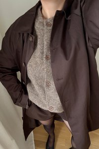 Brūns ūdensizturīgs garais mētelis virs beidzot adīta beigās džempera ar brūniem pogām. Melni šorti un zeķes piešķir ansamblim pabeigtību, uz gaismas fona.