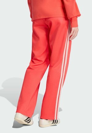 Czerwone sportowe spodnie o luźnym kroju, z białymi paskami po bokach i miękką teksturą, zestawione z białymi butami w czarne paski.