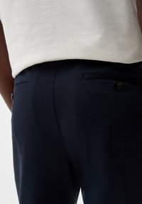 Námořnické látkové kalhoty s hladkým povrchem, které mají dvě zadní kapsy a detail s knoflíkem. Materiál se jeví jako lehký a přizpůsobený postavě.