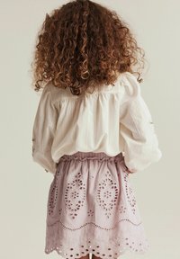 Weiße Bluse mit langen Ärmeln und gerafftem Rücken, kombiniert mit einem lila Rock, der filigrane Eyelet-Stickerei und einen gewellten Saum aufweist.