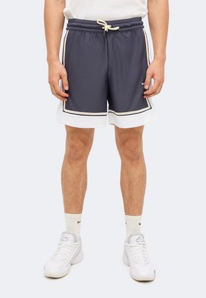 Le mannequin porte un short de sport gris foncé avec des bordures blanches et un cordon de serrage, des chaussettes blanches et des baskets blanches sur un fond uni.