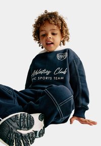 Navy sweatshirt en sweatpants set met witte accenten, voorzien van de teksten "Athletics Club" en "Epic Sports Team". Krullend haar is zichtbaar.