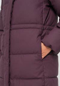 Pufferjakke i mørk lilla med en quiltet design, sidelommer og en glatt, matt overflate. Detaljene inkluderer sydd horisontale linjer.