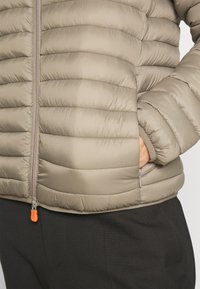 Beige puffer jacket with horizontal stitching, zippered front, and side pockets. Features an orange zipper pull accent.