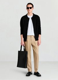 Cardigan noir, t-shirt blanc, pantalon beige et mocassins noirs. Tient un cabas noir à texture lisse et forme structurée. Des lunettes de soleil complètent le look.
